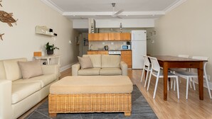 A living room with a sofa, ottoman, and dining table with chairs. A kitchen is visible in the background. - Caprice Court 6 (Cape Town)