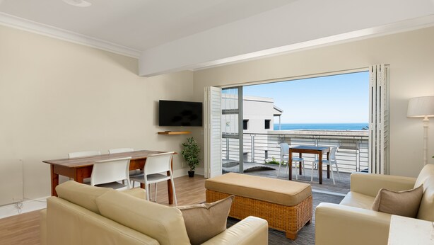 A modern living room with a sofa, a dining table, and a TV. There is a balcony with a view of the sea. - Caprice Court 6 (Cape Town)