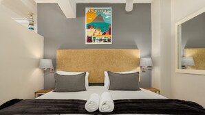 A hotel room with a bed, two bedside lamps, a headboard, a poster on the wall, and a mirror on the wall. - Caprice Court 6 (Cape Town)