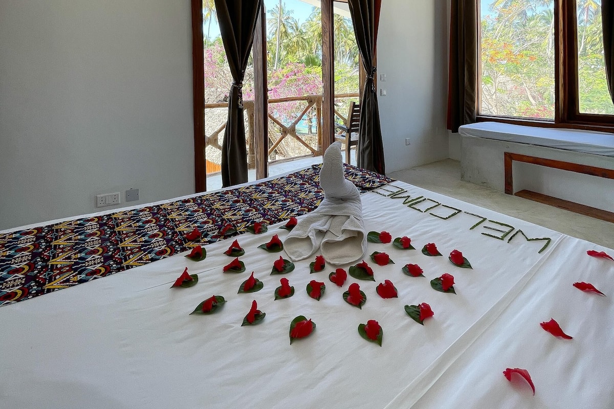 Quarto de hotel com uma cama, uma janela com cortinas, um banco junto à janela e vista para palmeiras e vegetação lá fora.