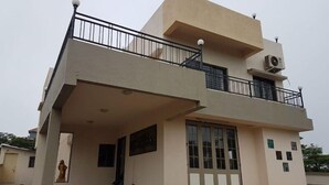 A two-story building with a balcony, a dining area with chairs and a table, and an air conditioning unit on the upper floor. - Vue Villa (Igatpuri)