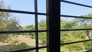 A window with a metal grid, through which a view of trees and a fence is visible. - Vue Villa (Igatpuri)