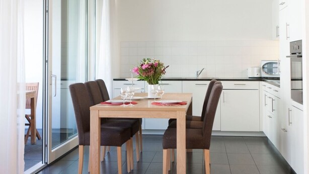 Une cuisine moderne avec des meubles blancs, une table à manger avec des chaises et une porte-fenêtre coulissante donnant sur un balcon.