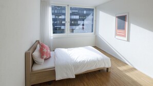 Une petite chambre avec un lit simple, un cadre en bois, du linge de lit blanc et une fenêtre donnant sur un immeuble.