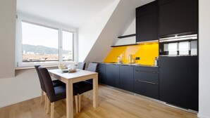 A modern kitchen with a dining area, featuring a wooden table and chairs, black cabinets, and a large window offering a city view.