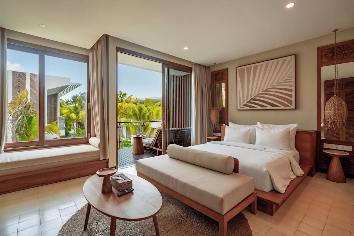 A bedroom with a large bed, a small table, a sofa, and a balcony with a view of greenery.
