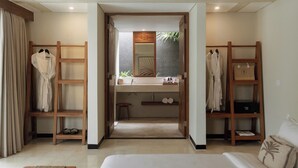 Habitación de hotel con cama, armario de madera, tocador con espejo y estante con toallas y artículos de aseo.