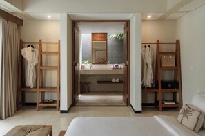 A hotel room with a bed, a wooden wardrobe, a vanity with a mirror, and a shelf with towels and toiletries.