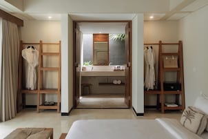 Habitación de hotel con cama, armario de madera, tocador con espejo y estante con toallas y artículos de aseo.