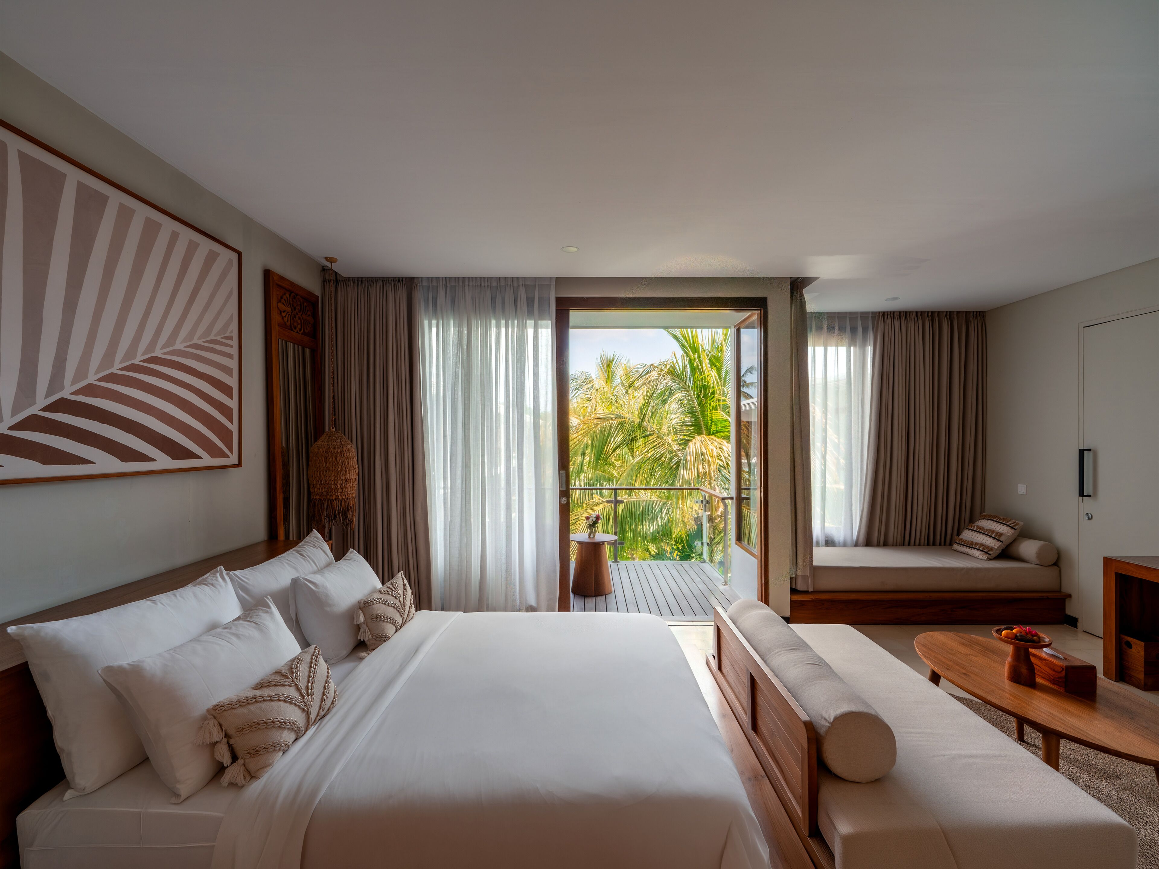 A hotel room with a large bed, a sofa, a small table, and a balcony with a view of palm trees.