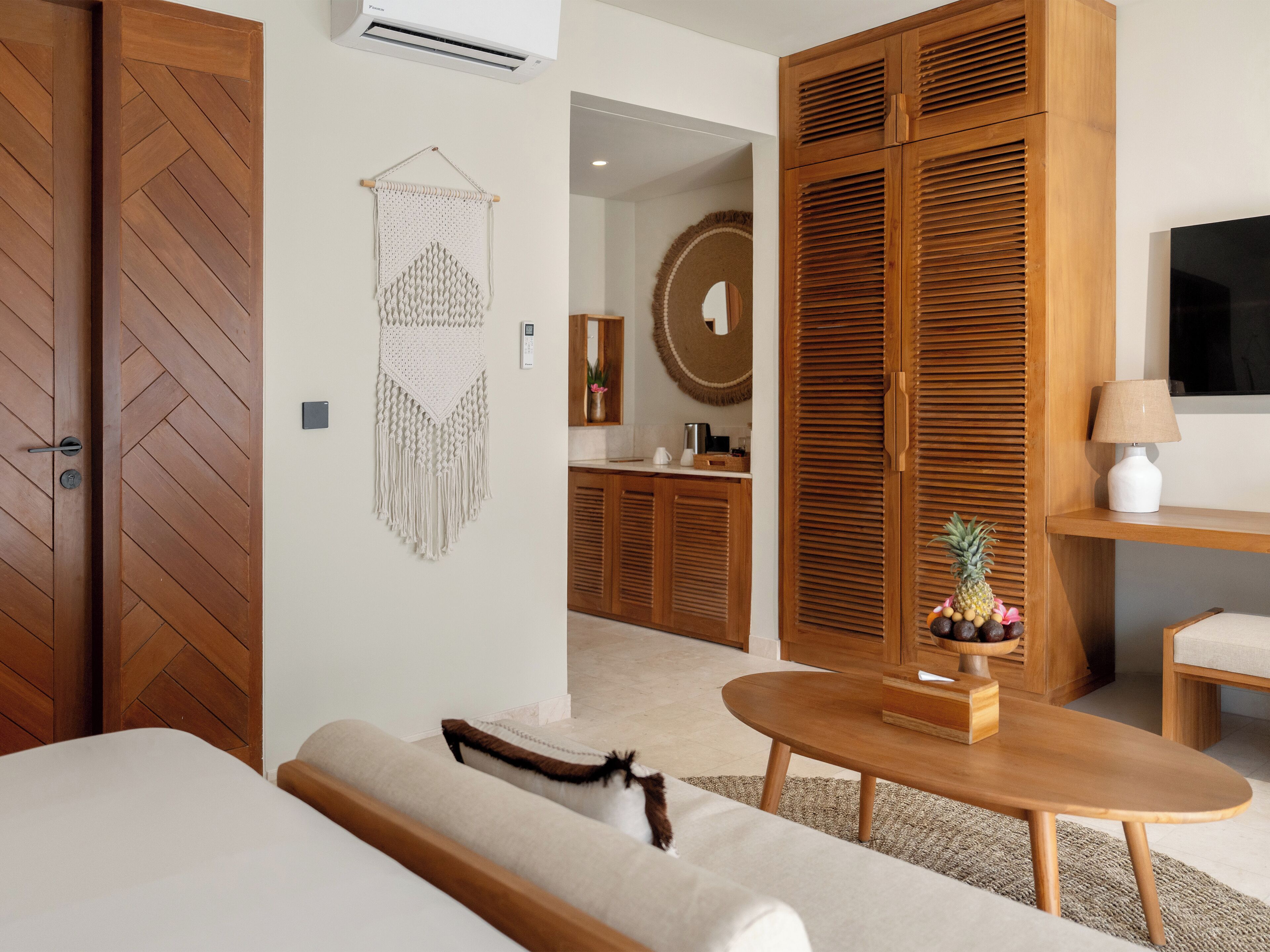 A hotel room with a herringbone-patterned door, a macrame wall hanging, a round mirror, a wooden wardrobe, a round table, a sofa, and a television on a stand.
