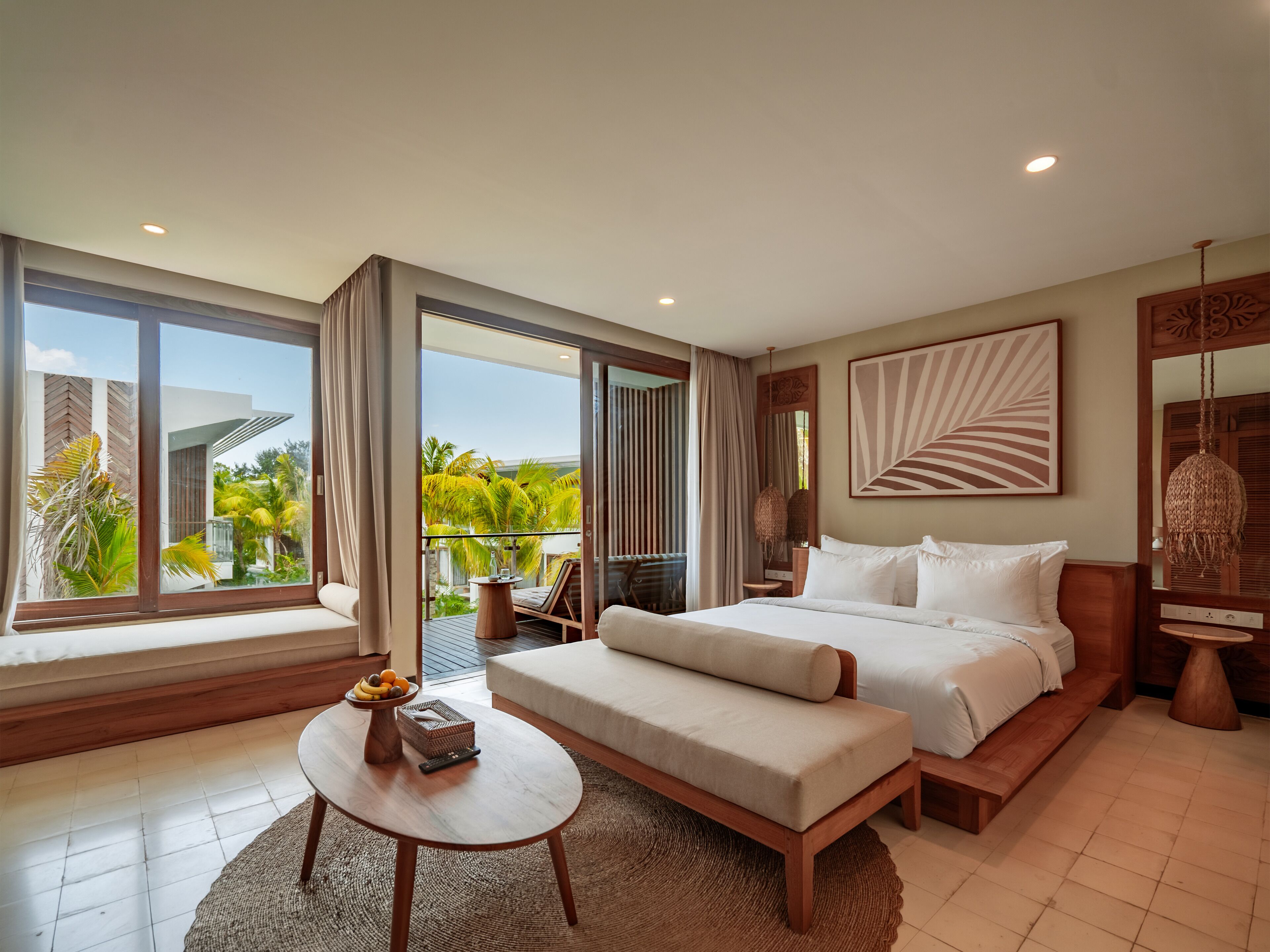 A modern hotel room with a large bed, a sofa, a small table with fruit, and a view of palm trees.