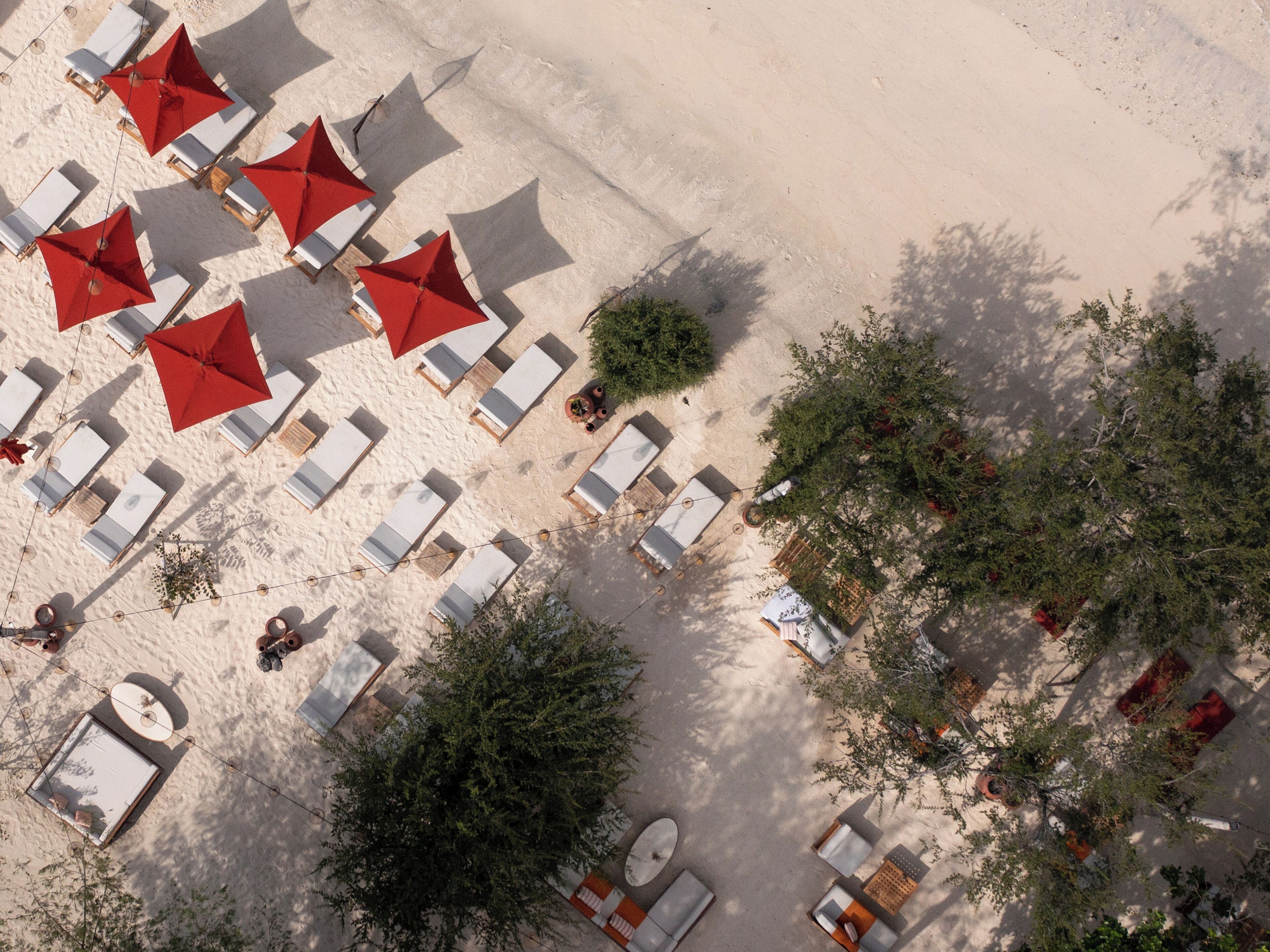 An aerial view of a beachside resort with sun loungers and red umbrellas, surrounded by greenery.