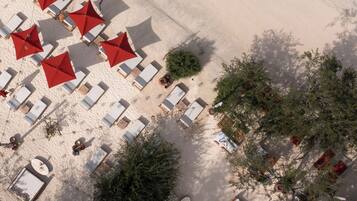 An aerial view of a beachside resort with sun loungers and red umbrellas, surrounded by greenery.