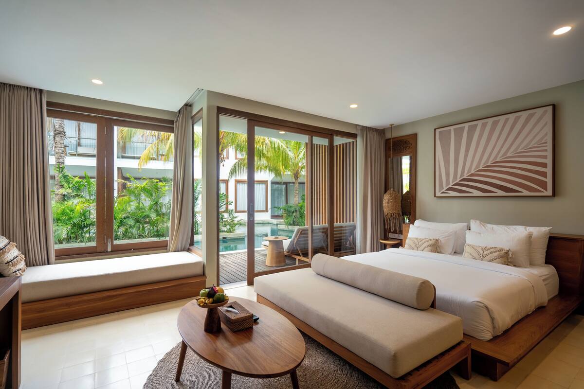 A modern bedroom with a large bed, a sofa, a small table with fruit, and a view of a pool and greenery.
