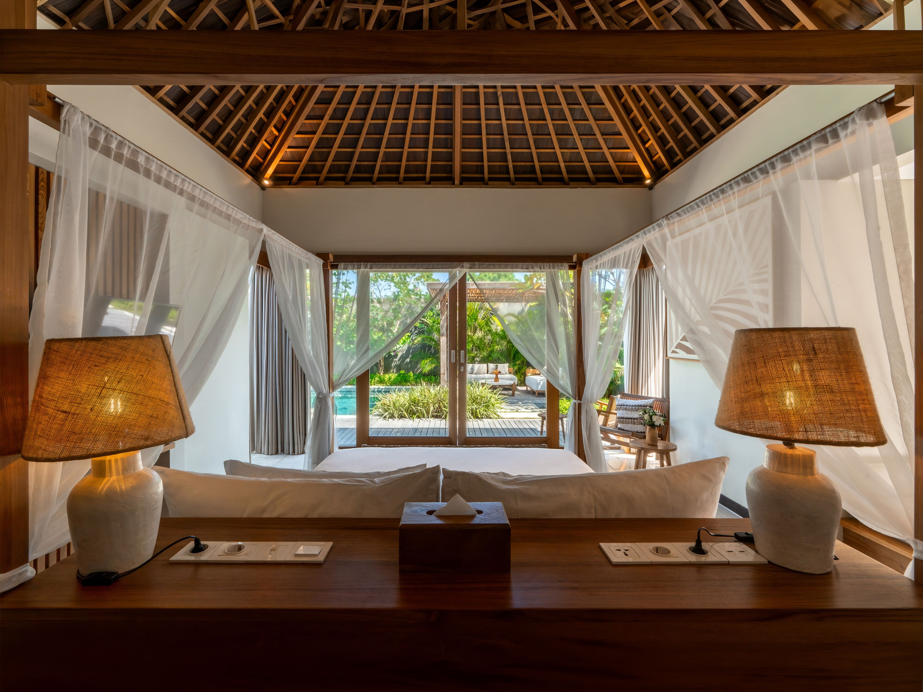 Una habitación con cama, lámparas de noche, mesita de noche y vistas a un área exterior con piscina y vegetación.