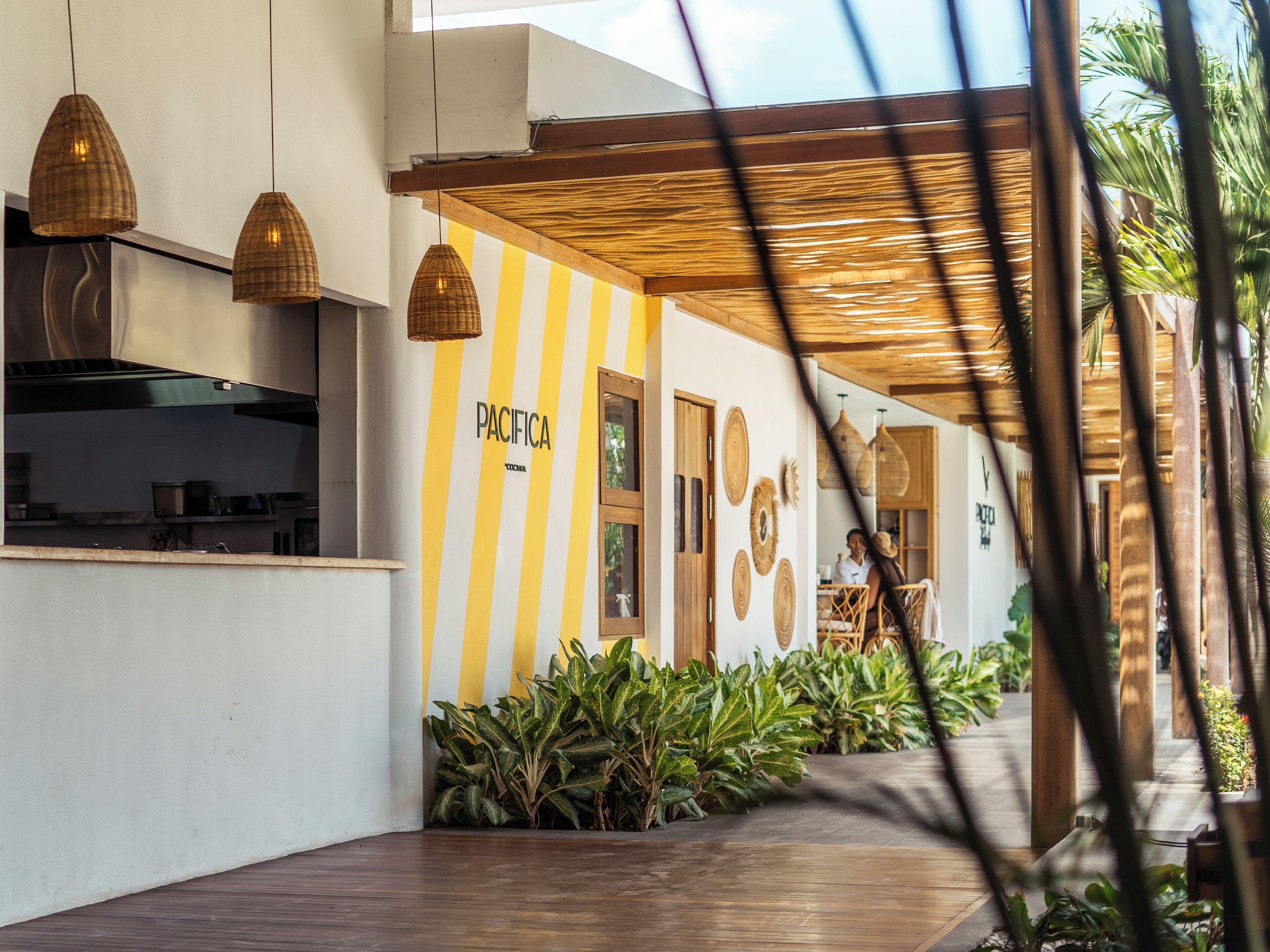 Un bâtiment moderne avec un mur rayé jaune et blanc, un espace cuisine et un coin salon extérieur abrité, agrémenté de plantes.