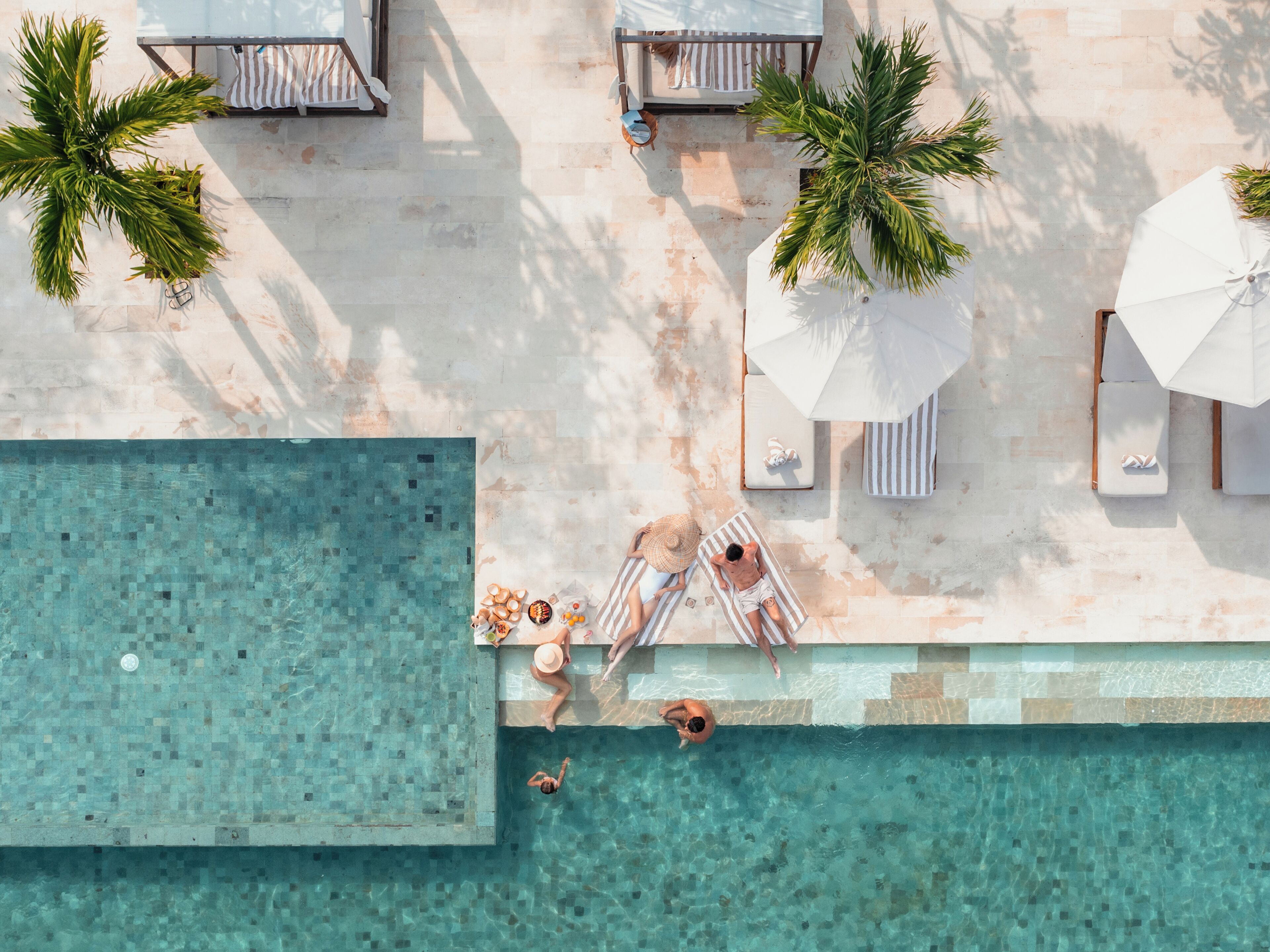 Vista aérea de una zona de piscina con sillones de descanso, sombrillas y palmeras.