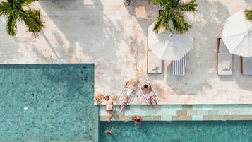Vista aérea de una zona de piscina con sillones de descanso, sombrillas y palmeras.