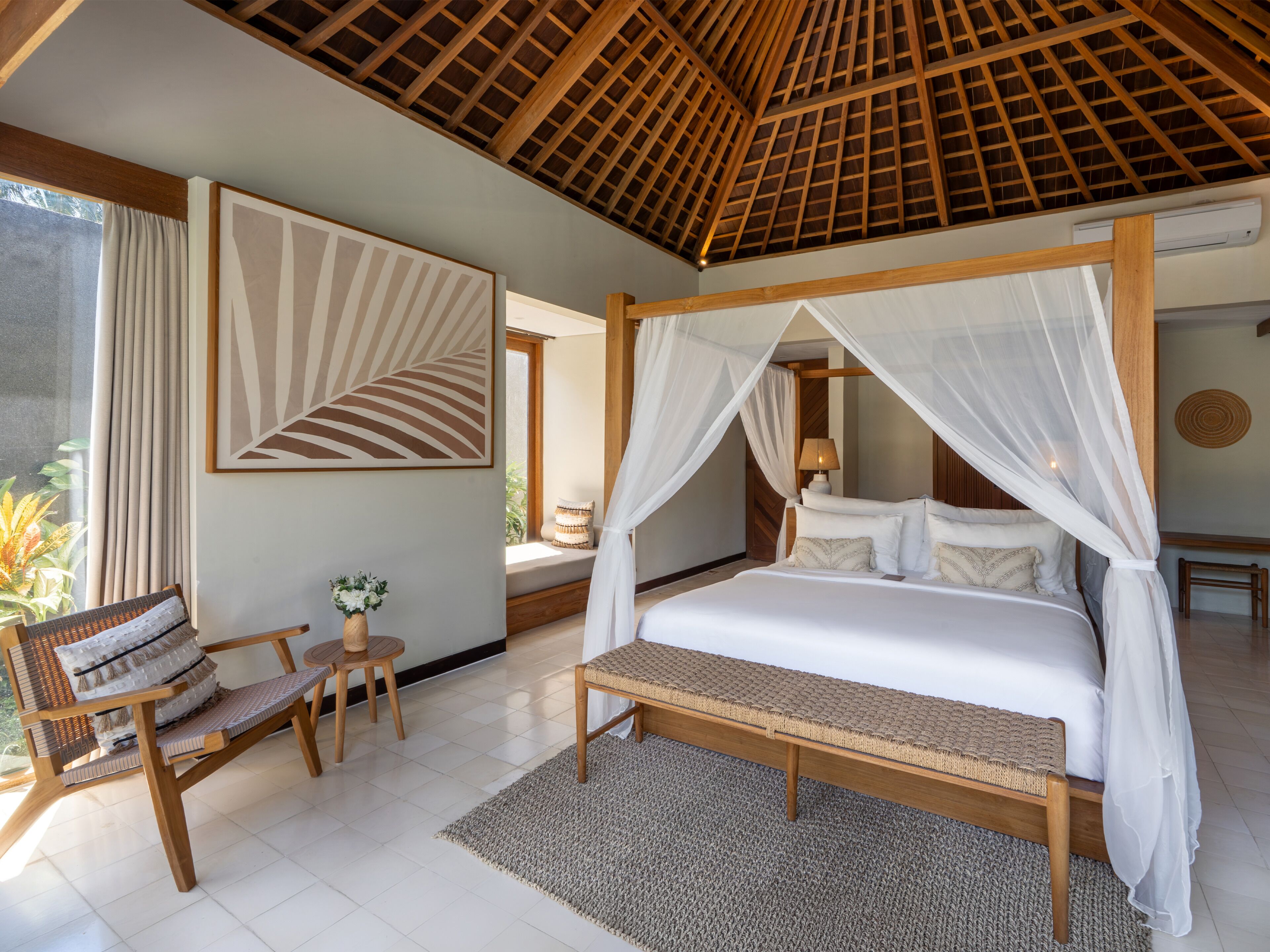 A bedroom with a canopy bed, a sitting area with a chair and table, and a large window with curtains.