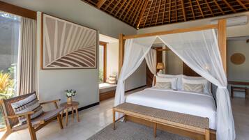 A bedroom with a canopy bed, a sitting area with a chair and table, and a large window with curtains.