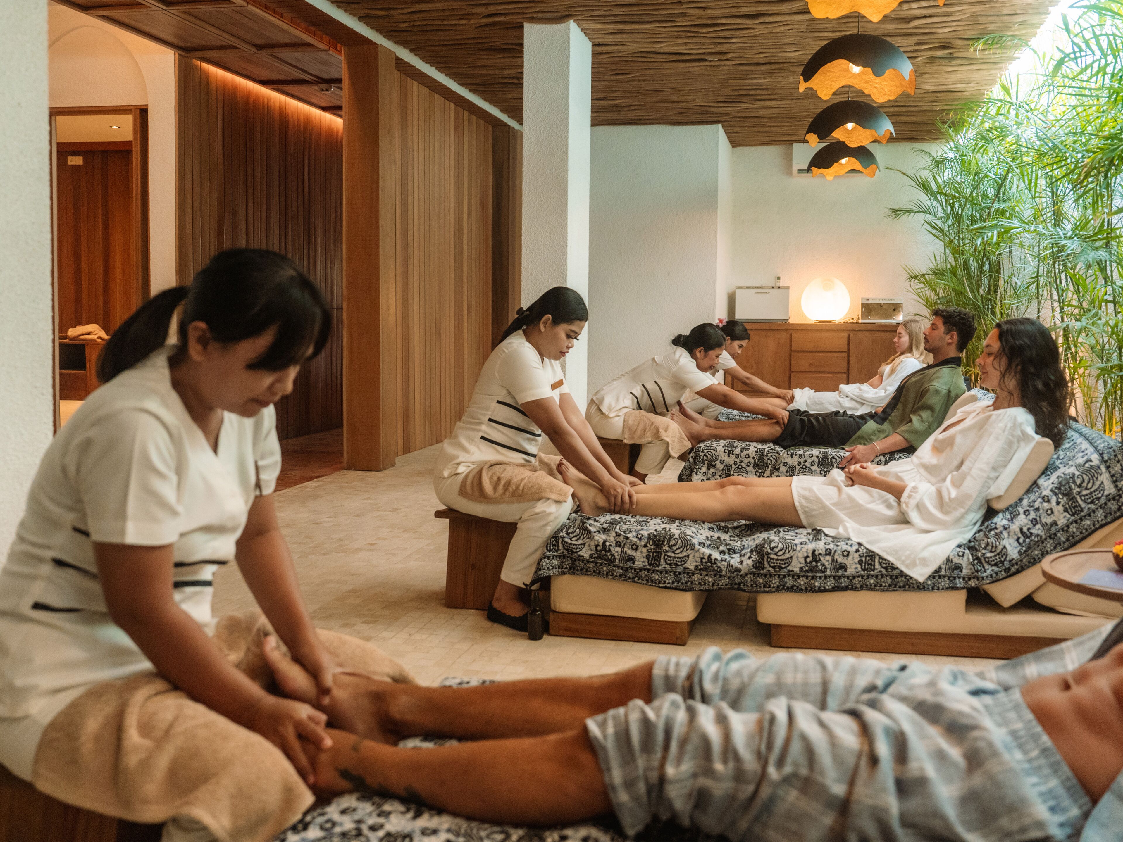 A massage therapy session with clients and therapists in a room with wooden paneling and a ceiling with hanging lamps.