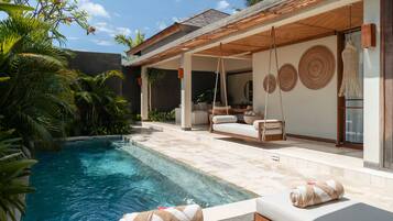 Un área junto a la piscina con una silla colgante, un sillón y un banco con toallas.
