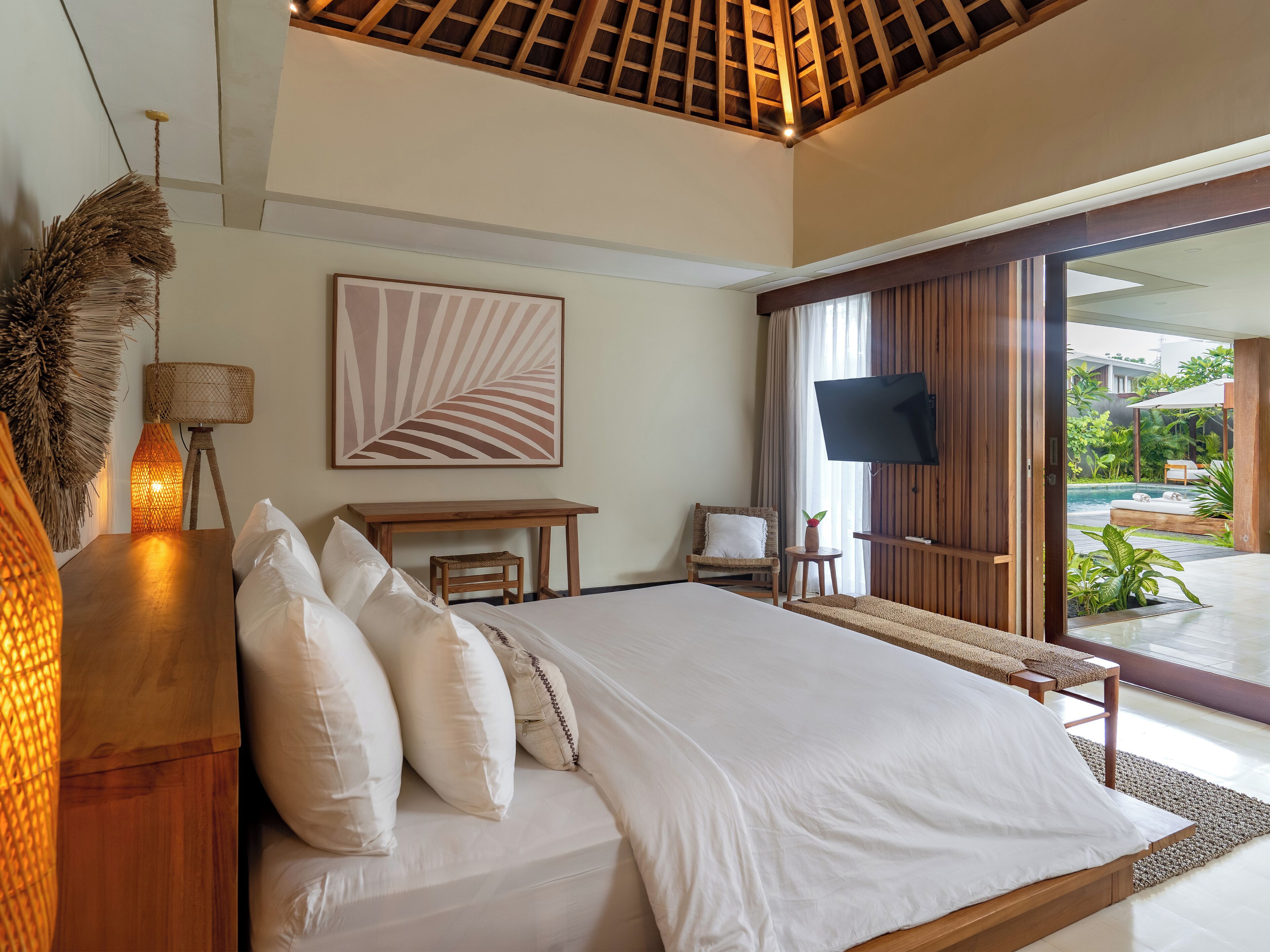 A bedroom with a large bed, a desk, a chair, a TV, and a view of an outdoor pool area.