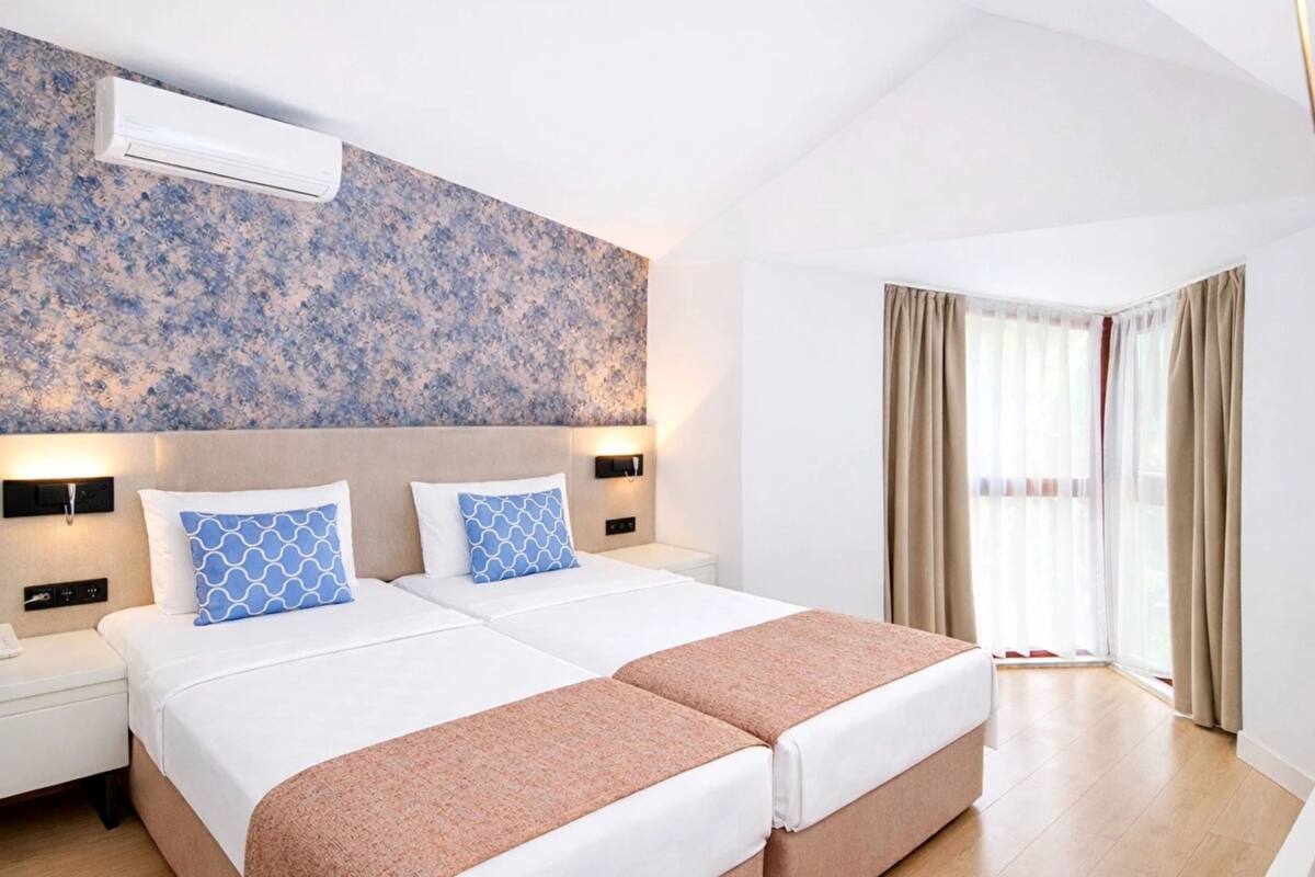 A hotel room with two beds, a large window with curtains, an air conditioning unit, and a wall-mounted light fixture.
