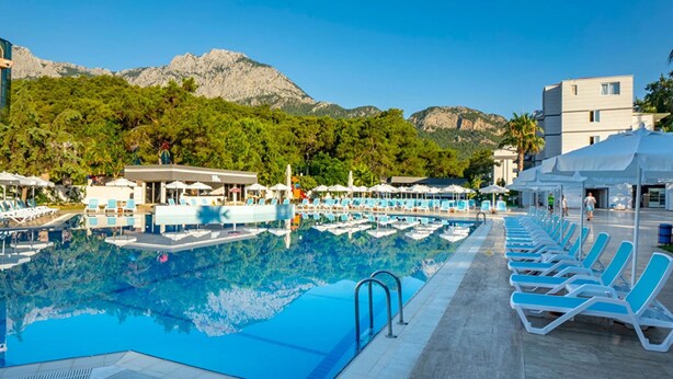 A swimming pool with lounge chairs and umbrellas, surrounded by buildings and mountains. - Alder Garden Resort - Ultra All Inclusive (Kemer)
