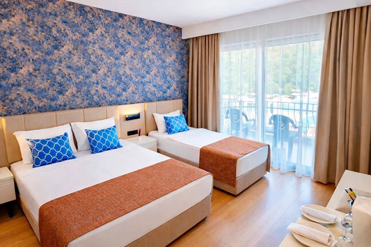 A hotel room with two beds, a large window with curtains, a balcony with chairs, and a table with a phone and glasses.