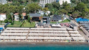 An aerial view of a beachside resort with multiple beach umbrellas and lounge chairs, a pool area, and surrounding buildings. - Alder Garden Resort - Ultra All Inclusive (Kemer)