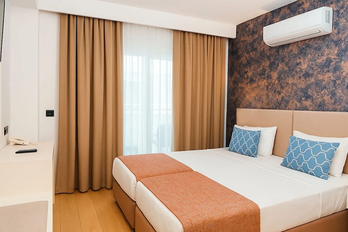 A hotel room with a large bed, beige curtains, a wall-mounted air conditioner, and a bedside table with a remote control.