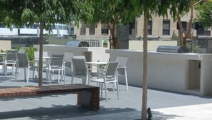 Un patio al aire libre con sillas y mesas blancas, un banco de madera y vistas a un edificio con fachada y ventanas blancas.