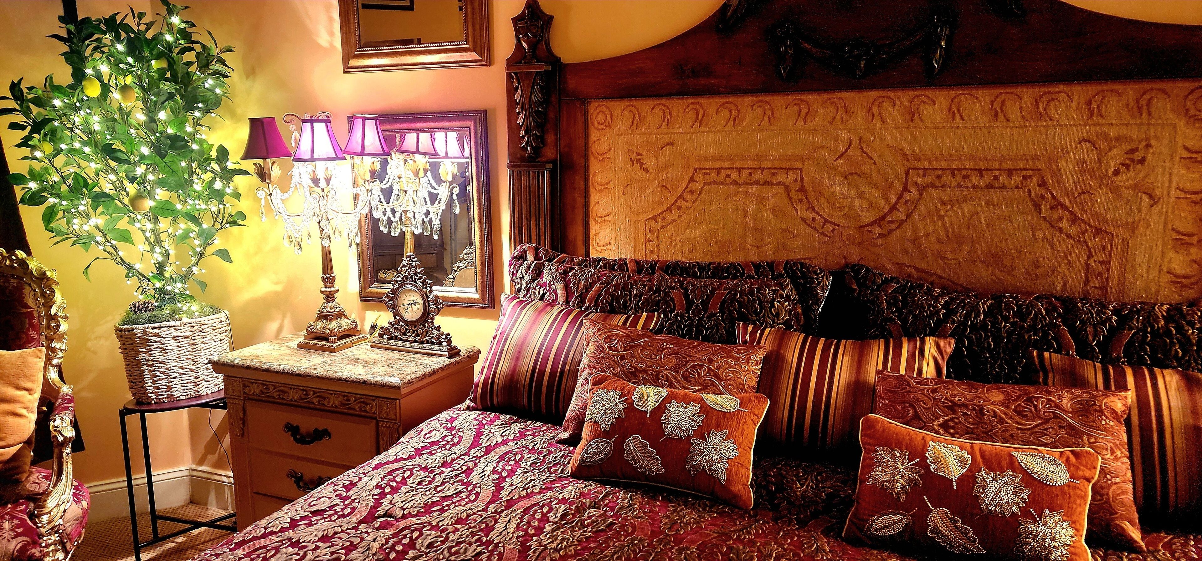 Una habitación tradicional con cama, cabecera ornamentada, planta en maceta, lámpara y espejo.