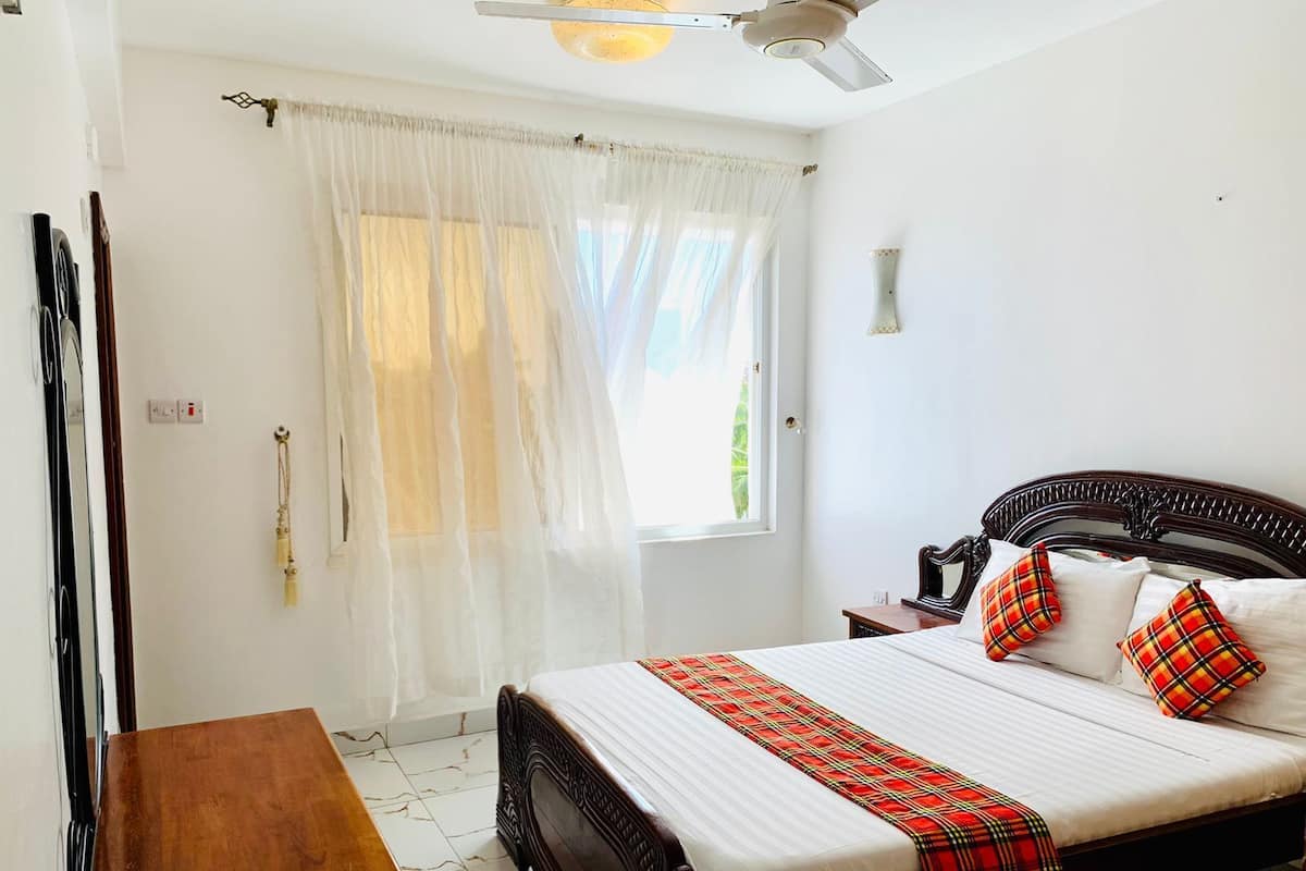 Un dormitorio con una cama grande, una mesita de noche de madera, un ventilador de techo y una ventana con cortinas translúcidas.