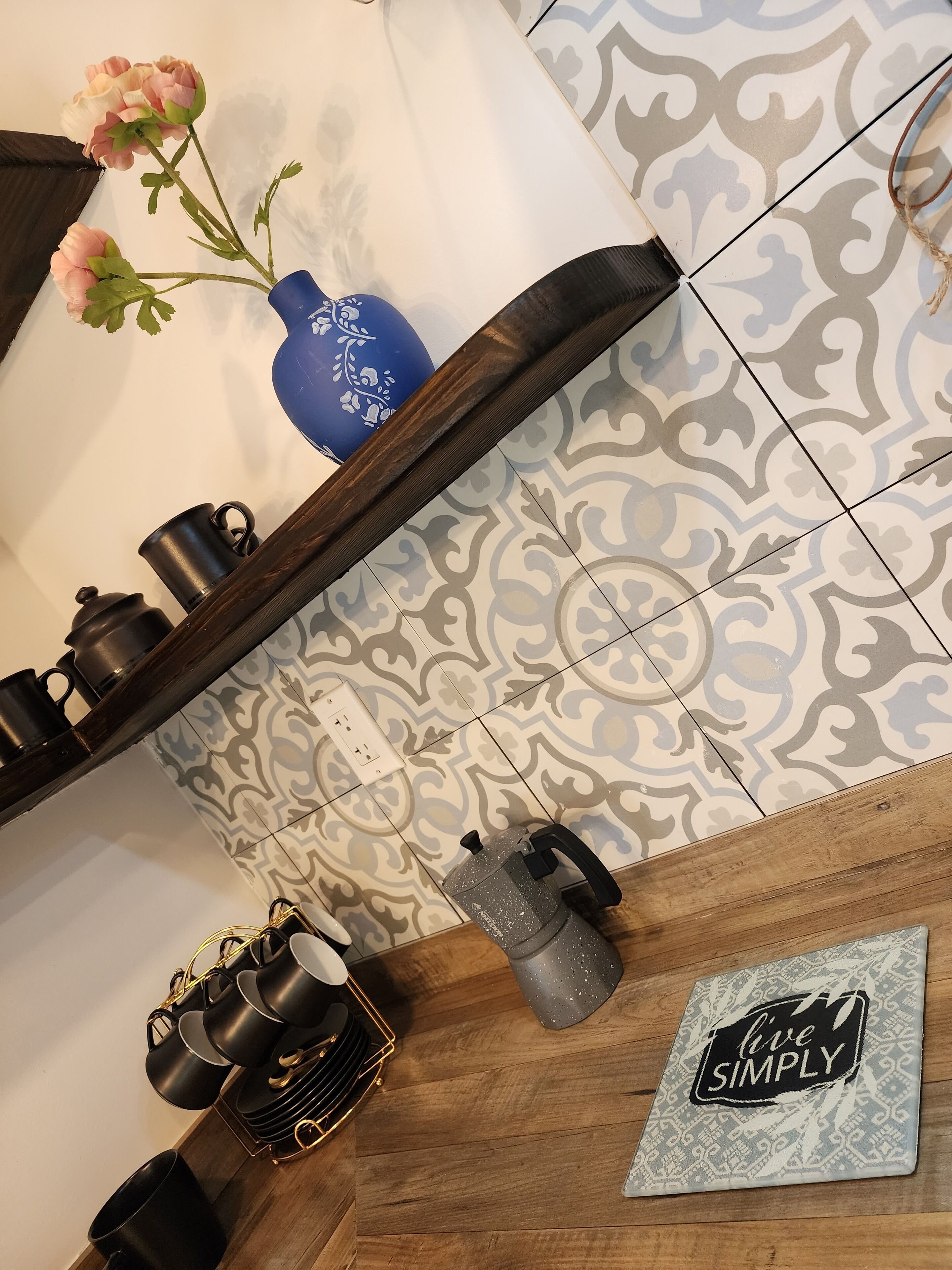 A kitchen with a wooden shelf holding black pots and a kettle, a blue vase with flowers, and a 'live simply' sign on the counter.