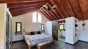 Een slaapkamer met een houten plafond, een bed met beddengoed met een patroon, een bankje en een schuifdeur naar een badkamer.