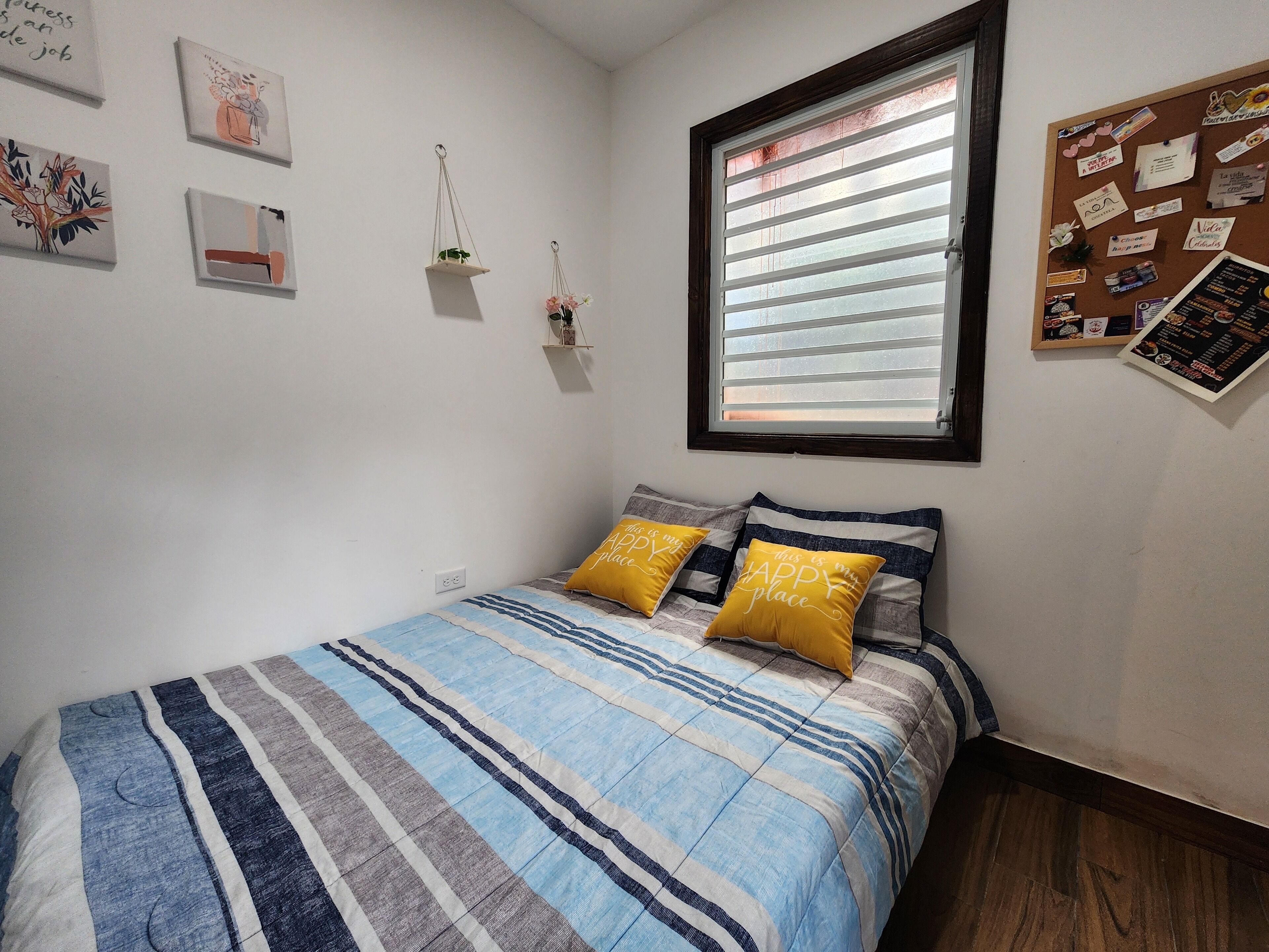Een slaapkamer met een bed, een raam en een prikbord aan de muur.