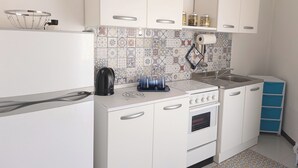 Una cocina compacta con gabinetes blancos, un salpicadero de azulejos, una tetera, un fregadero y una estufa.