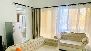 Un salon avec deux canapés beiges, un réfrigérateur blanc et une fenêtre ornée de rideaux légers.