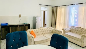 Un salon avec deux canapés de couleur crème, un fauteuil bleu et une table blanche. Il y a également une petite cuisine équipée d’un évier et d’un réfrigérateur.
