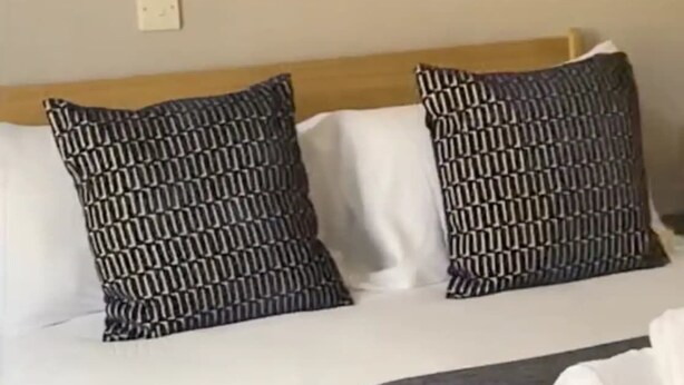 Un lit avec une couverture à motifs gris, des oreillers blancs et une tête de lit en bois.