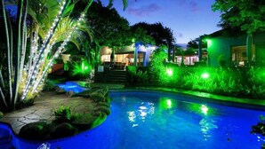 Vue nocturne d’un espace piscine avec des plantes illuminées et un bâtiment en arrière-plan.