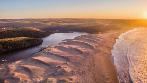 Une vue panoramique d’un paysage côtier avec une rivière, des dunes de sable et un ciel dégagé.