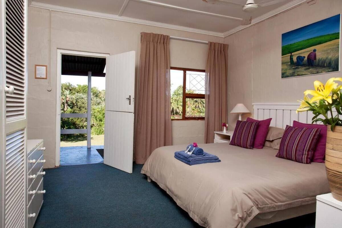 A bedroom with a bed, a nightstand, a lamp, a window with curtains, a painting, and a view of the outdoors.