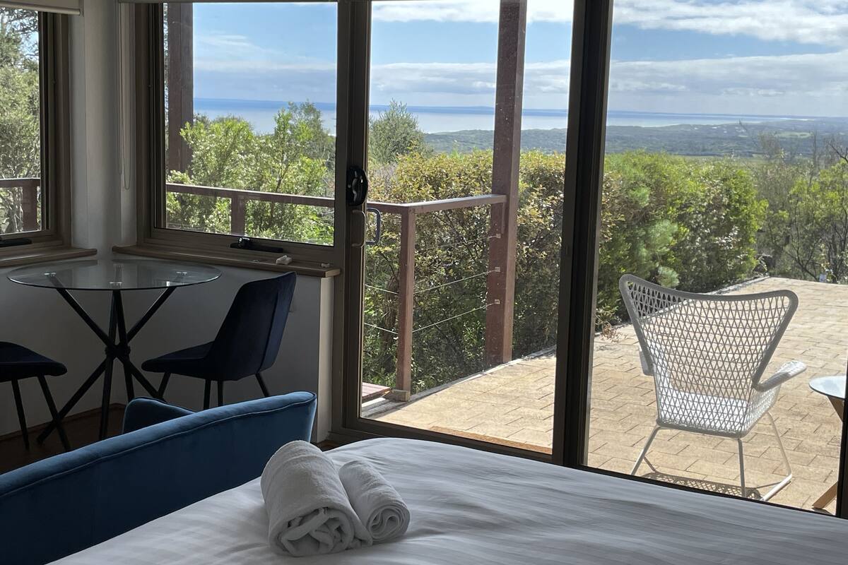 A room with a bed, a table, and chairs, a balcony with a view of greenery and a distant body of water.
