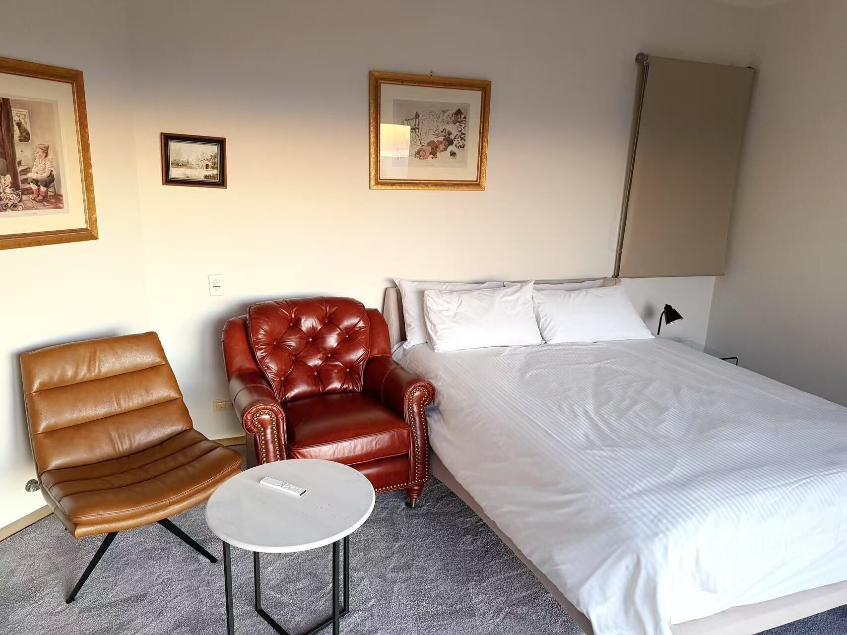 Una habitación de hotel con una cama, un sillón de cuero marrón, una mesita redonda y dos cuadros en la pared.