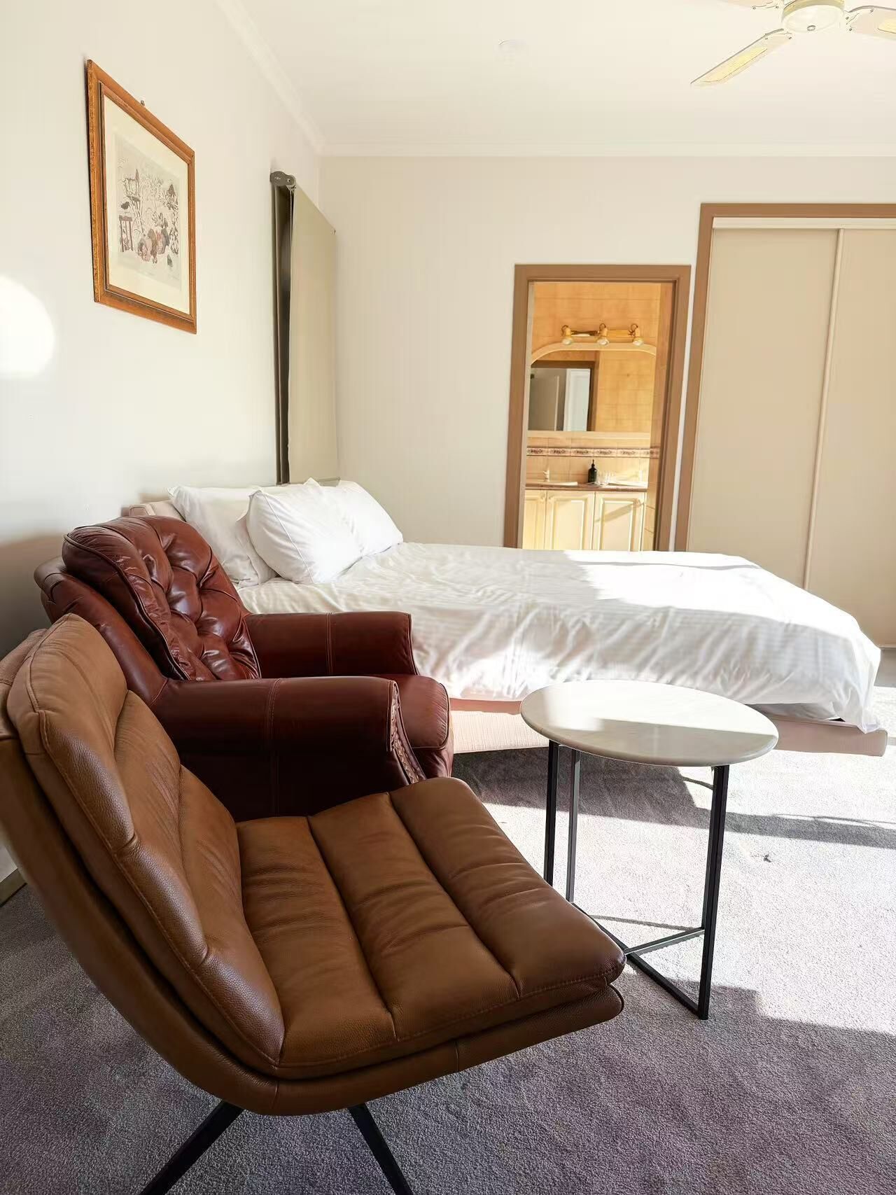 Habitació d'hotel amb un llit, una cadira de pell marró, una taula petita i un ventilador de sostre.
