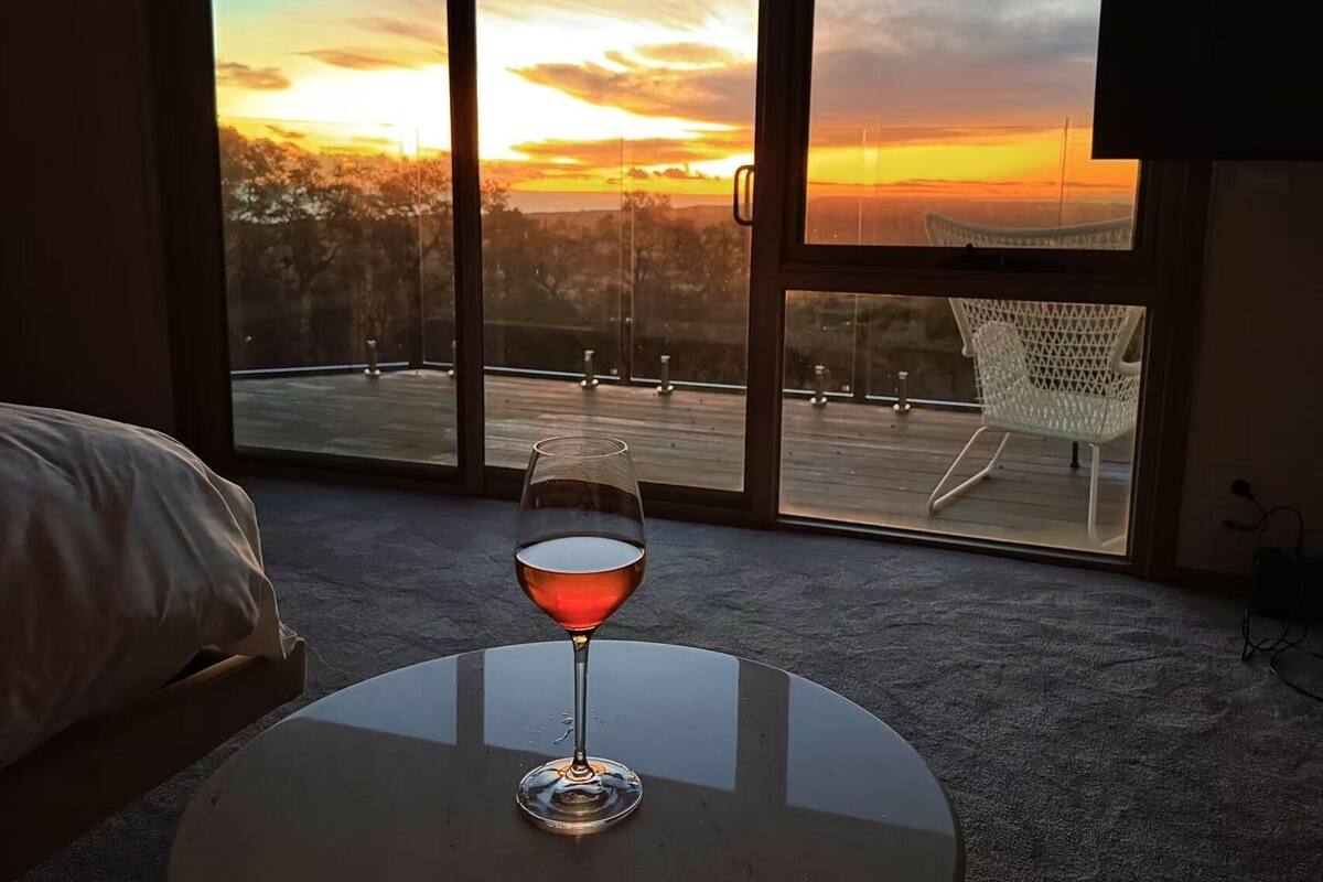 A room with a glass of red wine on a table, a bed, and a balcony with a chair.